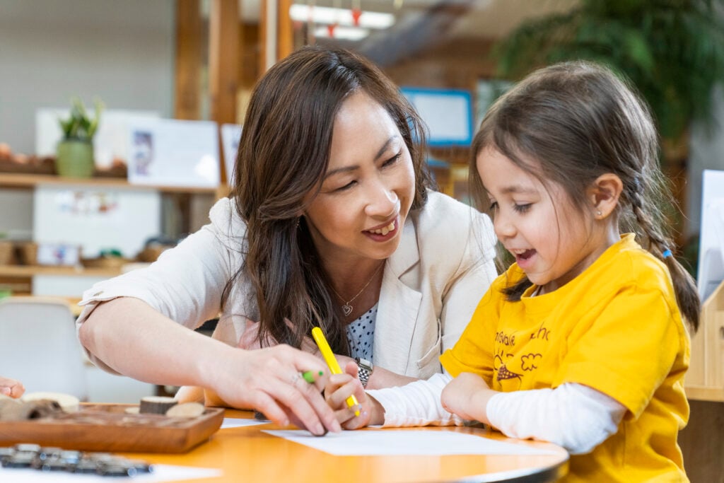 A picture of a young primary school student in a classroom with their teacher smiling as they learn. 